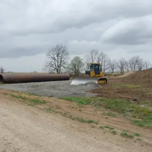 Dozer spreading rock for the equipment access pad, with large-diameter pipe staged at the edge