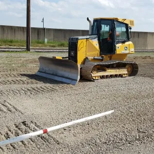Tracked dozer with the blade lowered on the gravel platform alongside the floodwall and rail, a stringline and grade pole set in the foreground