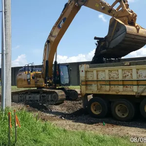 Tracked excavator loading excavated material into a tandem dump truck while the crew watches from the floodwall side