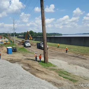 Elevated view of the work corridor between the floodwall and adjacent businesses, with staged equipment, dump trucks and crews and the Mississippi River beyond the wall