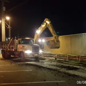 Excavator working beside the floodwall at night under street lighting with a dump truck positioned alongside