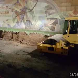Smooth-drum vibratory roller compacting the excavated strip at night in front of the painted floodwall