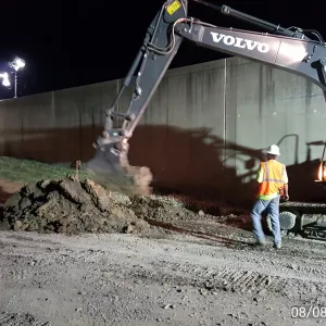 Excavator casting spoil into a pile at the toe of the floodwall at night while a worker looks on