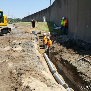Open trench at the toe of the floodwall with new pipe laid in the bottom, a crew working at a precast structure and drain rock placed against the wall