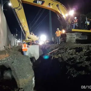Worker guiding a precast concrete structure into the trench at night as an excavator holds the load