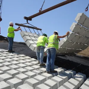 Crane and spreader beam lowering a mat of articulated concrete block onto geotextile while three workers guide it into position