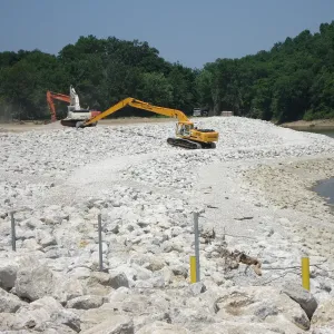 Long-reach excavator working large riprap along the channel bank with water at right and fence posts set through the stone in the foreground