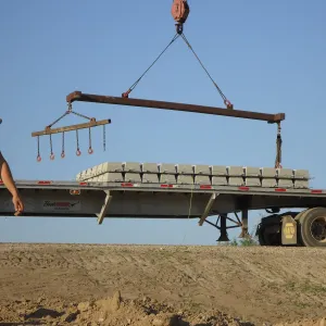 Worker walking past a flatbed trailer loaded with articulated concrete block mats as a crane spreader beam and lifting bar hang overhead