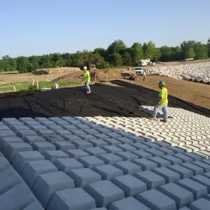 Workers placing articulated concrete block over geotextile on a graded slope, with stockpiled riprap lining the channel beyond