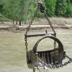 Close view of a perforated clamshell bucket suspended above muddy flowing water in front of an eroded, debris-strewn bank