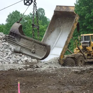 Clamshell bucket swinging over a crushed-rock stockpile as an articulated dump truck dumps its load alongside