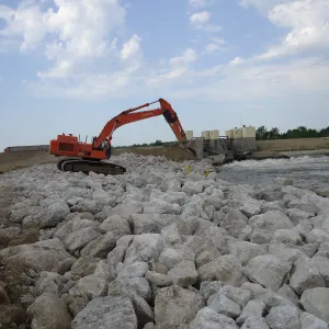 Excavator placing large riprap on the bank next to the dam outlet gates with water discharging through them