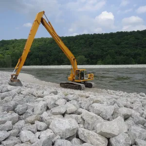 Long-reach excavator sitting on a riprap-covered bank, reaching toward the water with rock revetment continuing along the far side