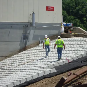 Two workers walking across placed articulated concrete block where it meets the concrete wall of the outlet structure, below a red warning sign