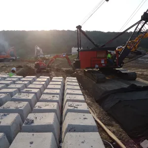 Crawler crane and excavators working in low sun beside rows of concrete block and geotextile laid into a trench