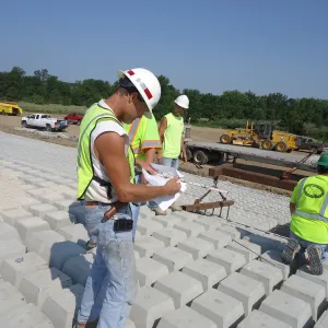 Workers checking a drawing and a level on placed concrete block, with a flatbed of blocks and a motor grader on the bench behind