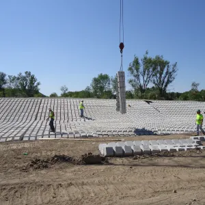 Crane lowering a vertical bundle of concrete blocks onto a broad completed field of block revetment as workers walk across it