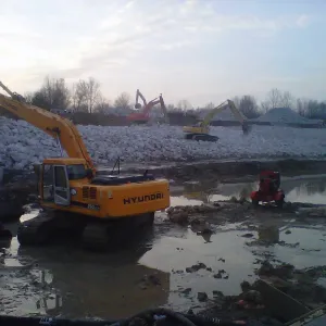 Several excavators working large riprap stockpiles at dusk beside a muddy, water-pooled work area with a pump set at the edge