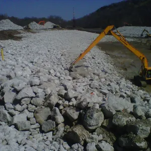 Long-reach excavator spreading large riprap across a wide stone-covered channel bottom, bare trees on the hillsides