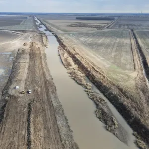Aerial of the channel with crew trucks and a dozer working the top bank near a road crossing