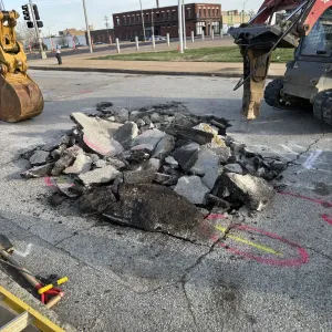 Broken asphalt and concrete piled in a saw-cut pavement opening at a signalized intersection, with an excavator bucket at left and a skid steer carrying a hydraulic breaker at right
