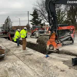 Skid steer with a hydraulic plate compactor working alongside a narrow open trench in a concrete street, with two workers at the trench edge and a second excavator staged behind