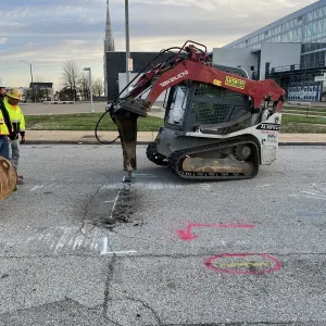 Tracked skid steer with a hydraulic breaker cutting through asphalt along a painted cut line, with utility locate markings on the pavement