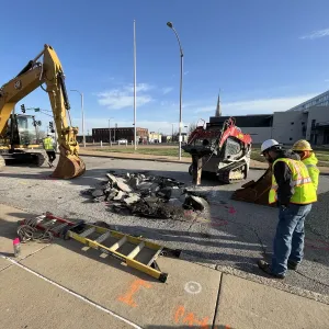 Crew and equipment around a broken-out pavement opening in a wide street, with an excavator staged at left, a skid steer at right, and marking paint on the sidewalk