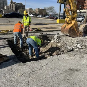 Three workers hand-digging in a shallow pavement opening in a street, with an excavator bucket and thumb positioned at right and a building main-entrance canopy in the background