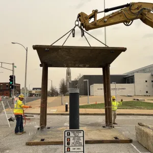 Excavator lowering a steel road plate onto a supported frame in a street, with two workers tending tag lines and a school building in the background
