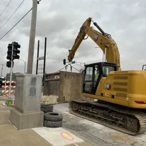 Excavator rigged with chains to a precast concrete structure section staged at the curb, with an electrical cabinet and orange barricades in the foreground