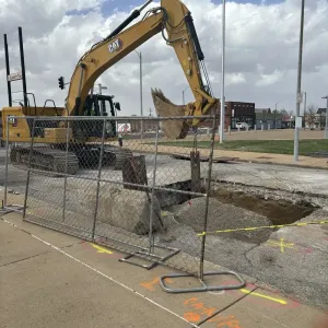 Excavator working behind chain-link fence panels at an open pavement excavation, with concrete blocks and timber staged at the hole edge and locate markings on the sidewalk