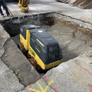 Walk-behind trench roller compacting the bottom of a wide pavement excavation, with an excavator arm above and timber sheeting stacked at the curb
