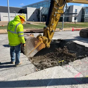 Worker guiding an excavator bucket as it digs a pavement opening in front of a school building on North Grand Boulevard