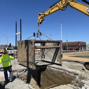 Excavator lifting a steel trench box on chains above an open excavation, with a worker on a tag line and a utility locating truck parked in the background
