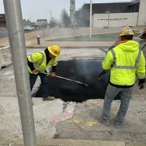 Two workers raking and screeding hot asphalt into a pavement patch, with steam rising and the school building behind