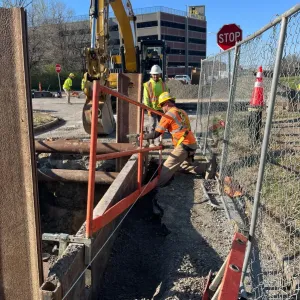 Worker in a fall-arrest harness setting bracing at the top of a shored trench with steel sheeting and pipe struts, with an excavator behind
