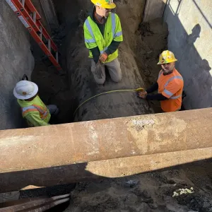 Three workers in a shored trench measuring the outside of a large-diameter pipe with a tape, with steel struts spanning the trench above them