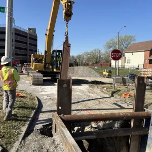 Excavator lifting a steel soldier beam on a rigging plate beside a shored trench, with a worker spotting at left and an aggregate stockpile behind
