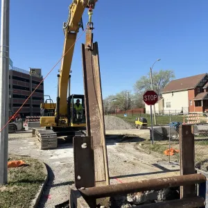 Steel soldier beam suspended from an excavator above a shored trench, with a residential street, stop sign, and parking garage in the background