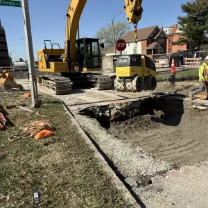 Excavator and walk-behind trench roller beside a partially backfilled excavation at Finney Avenue, with crushed stone bedding placed along the trench bottom