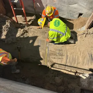 Three workers measuring and hand-digging around a large-diameter pipe inside a steel-plate shored excavation