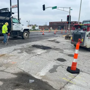 Worker with a shovel at the tailgate of a dump truck on a fresh asphalt patch at Bell Avenue and Grand Boulevard, with channelizer drums closing a lane and a skid steer at right