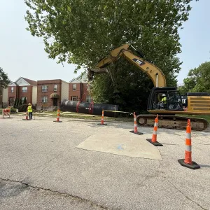 Excavator carrying a slung section of large-diameter pipe across a residential street inside a coned-off work zone, with brick two-family houses behind