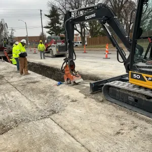 Compact excavator with a hydraulic plate compactor working the edge of a narrow trench in a concrete street, with workers observing and a skid steer behind