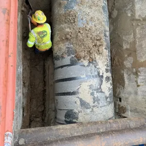 Worker hand-digging in a narrow shored trench alongside a large-diameter cement-lined pipe, with steel struts crossing above