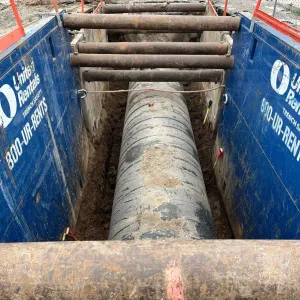 Large-diameter cement-lined pipe laid in the bottom of a shielded trench with rows of steel struts above, viewed down the trench line