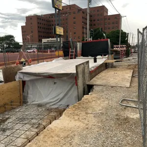 Tarped concrete formwork and reinforcing mesh in a fenced work corridor, with the Veterans Affairs Medical Center building across the street