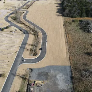 Curb-and-gutter roadway threading the expansion, with a gravel staging area and excavator at the work face