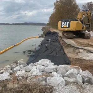 Geotextile fabric laid on the graded bank ahead of stone placement, with the turbidity barrier deployed offshore